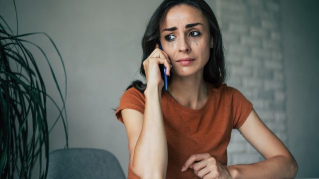 Sad, frustrated young brunette woman is crying with smartphone in hands while she sitting on the chair at apartment