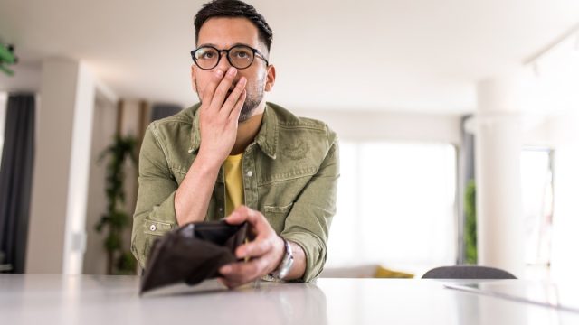 Young man sitting at home with an empty wallet. He is left without money and thinking what to do next.