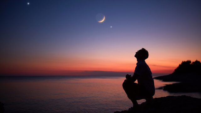 The silhouette of someone standing by the ocean and looking up at the moon and planets in the night sky at dusk