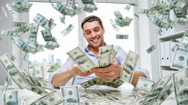 A rich young man sitting at his desk counting money with it flying all around him.