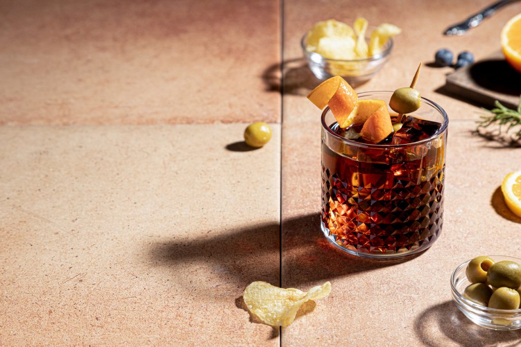 A closeup of a vermouth cocktail with an orange zest and green olive garnish, surrounded by potato chips