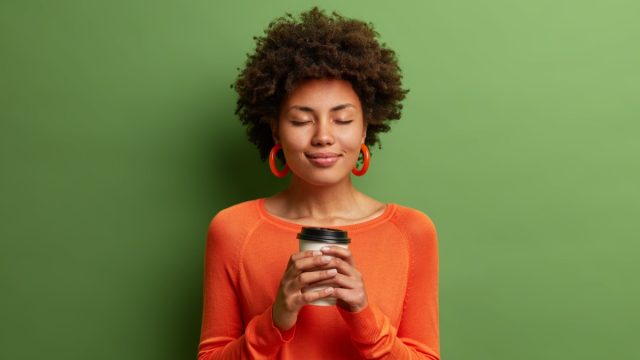 Studio shot of satisfied dark skinned girl with Afro hair holds paper cup of hot coffee, closes eyes, wears orange jumper, green background. Beautiful young woman enjoys aromatic cappuccino.