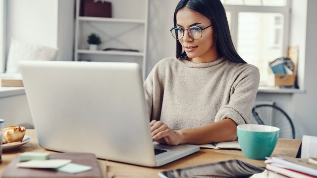 A young woman with long brown hair wearing blue glasses and a beige sweater is working on her laptop smiling.