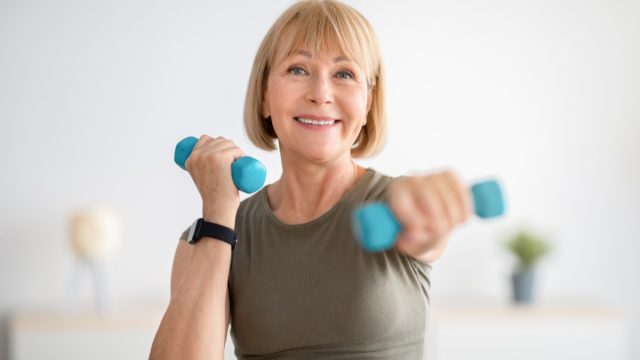 Strong senior woman doing exercises with dumbbells indoors.