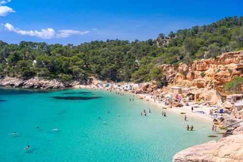 Wide-angle view of Cala Saladeta, a few steps from Cala Salada, on the western coast of Ibiza, renowned for its extremely transparent waters, pearly sands and a scenic group of fishermen’s huts (locally known as casitas varadero). Bathers engaged in different activities, the bright light of a summer noon, picturesque clouds, colourful cliffs, lush vegetation. 