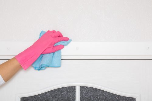 Young adult woman hand in pink rubber protective glove holding blue dry rag and wiping dust from white top frame of wooden door in room. 