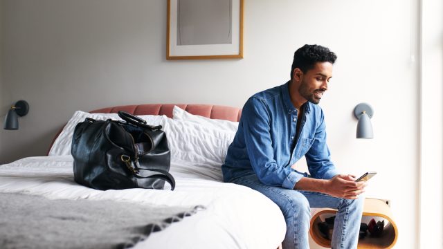 Male guest in boutique hotel sitting on edge of bed checking emails and social media on mobile phone in the morning