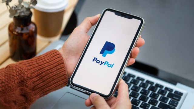 A close up of a smartphone with the PayPal logo on its screen and a computer in the background