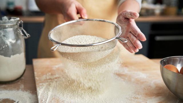 A close up of a person sifting flour over a cutting board in a kitchen