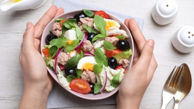 Woman holding bowl of delicious salad with canned tuna at white wooden table, top view