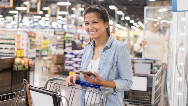 A woman smiling while pushing her cart through a warehouse bulk goods store and holding her smartphone