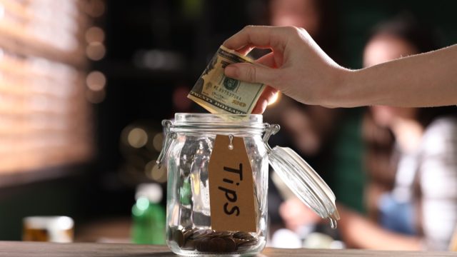Woman putting tips into glass jar on wooden table indoors, closeup