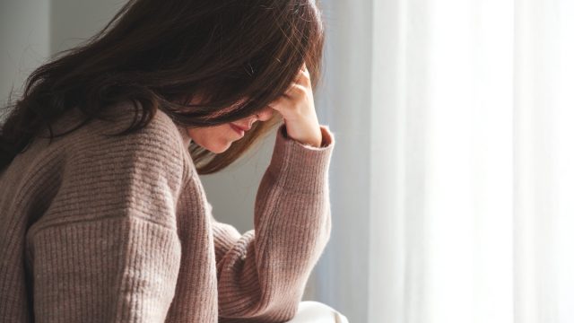 Closeup of a woman wearing a beige sweater sitting with her head in her hand and her long brown hair covering her face