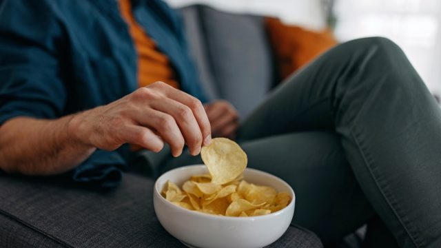 Young unrecognizable man at home, sitting on sofa and taking a potato chip from a bowl placed on a sofa