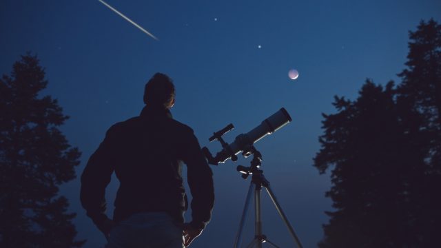 A stargazer standing next to a telescope and looking up at the sky as a meteor shoots overhead