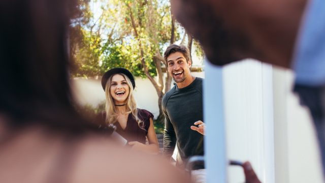 Excited couple at entrance door with wine bottle. People attending friend's housewarming party.