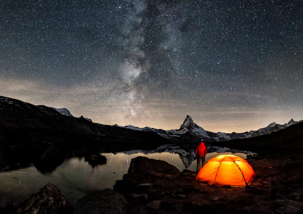 A person standing next to an illuminated tent looking up at stars in the night sky.