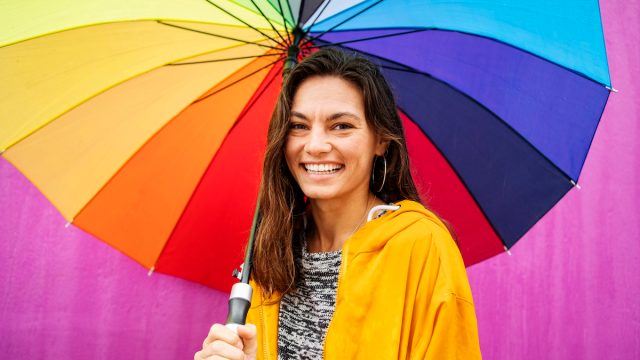 Portrait of a smiling young woman standing outside in the rain and holding a large rainbow colored umbrella