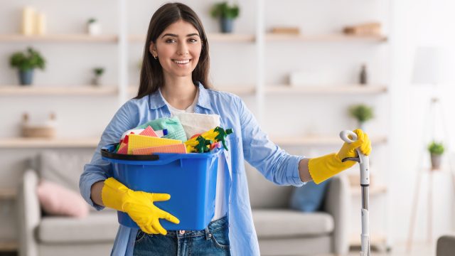 Portrait of smiling millennial lady holding bucket with cleaning supplies and mop, posing and looking at camera standing in living room.