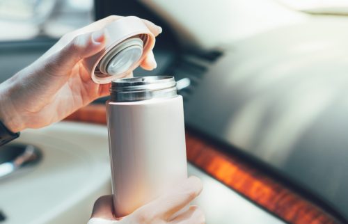 Closeup, Woman’s hands hold insulated reusable water bottle in car instead of single use plastic cup, open lid to drink her coffee. Eco friendly, zero waste and green living lifestyle.