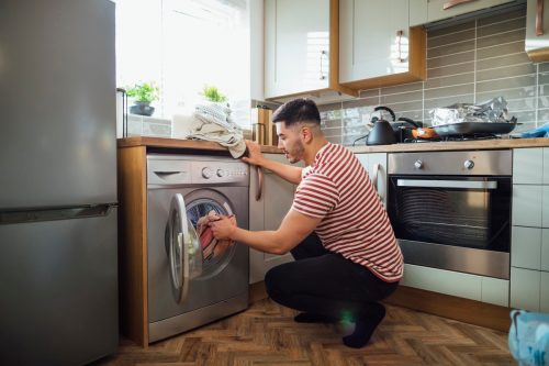 Man crouching down to put washing into his washing machine in his kitchen. He is in the Northeast of England.
