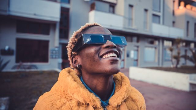 A person wearing an orange coat and protective solar glasses looking up into the sky with a smile