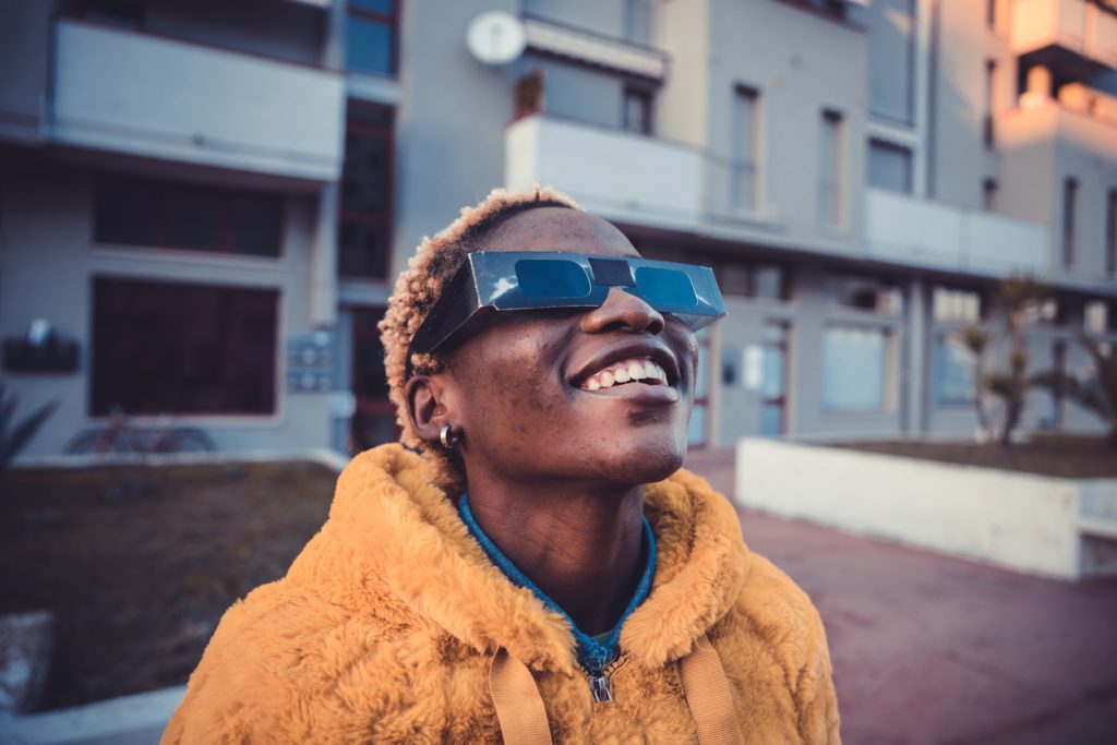 A person wearing an orange coat and protective solar glasses looking up into the sky with a smile