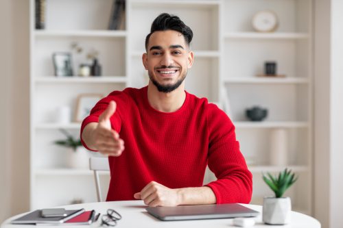 A smiling young man sitting in his office wearing a red sweater with his arm outstretched for a handshake