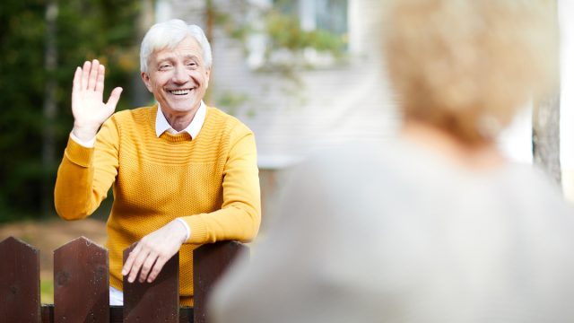 Cheerful and friendly mature man waving hand to his neighbour while standing by fence