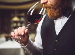 A close up of a person with a beard sniffing from a glass of red wine