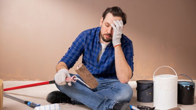 An upset-looking man sitting on the floor with his head in his hands while surrounded by painting supplies