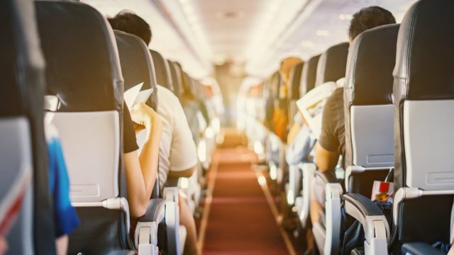 Interior of airplane with passengers sitting on seats and stewardess walking the aisle in background.
