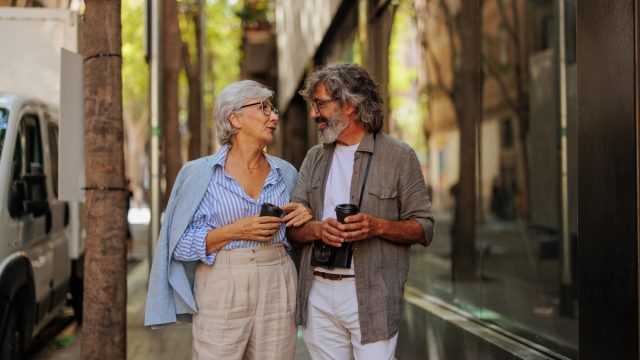 A cheerful senior couple is outside in the city walking on the street, socializing and drinking coffee.