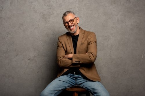 bearded businessman in glasses looking at camera and sitting with crossed arms on grey
