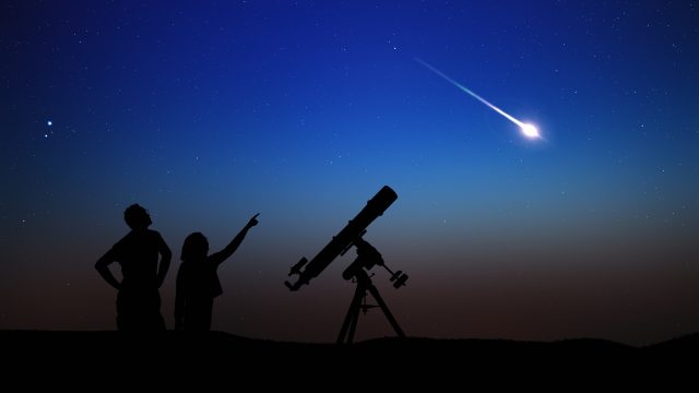 A silhouette of a parent and child watching a shooting star next to their telescope