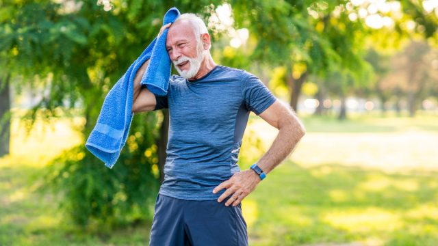 Senior man with white hair and beard wearing blue workout clothes takes a break while exercising outside on a hot day, wiping his face with a blue towel