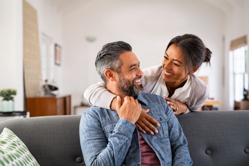 woman smiling at her husband and hugging him from behind the couch