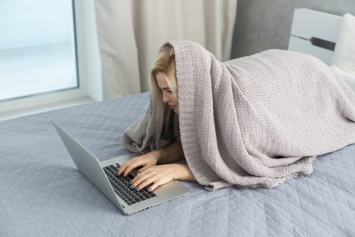 woman writing funny 'out of office' messages from bed while hiding under a blanket