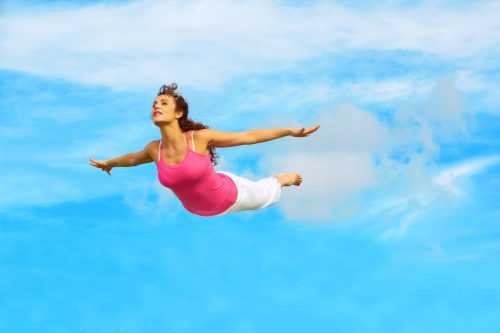 Full length of a sporty female flying in air with hands outstretched against sky.