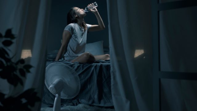 Young woman suffering from insomnia and heat, she is sitting on the bed and drinking fresh water with a fan blowing on her