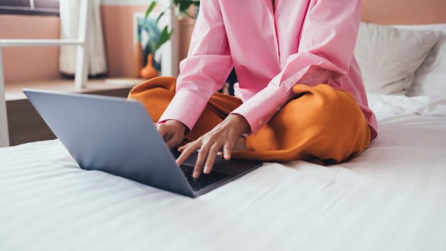 A woman sitting on a bed and typing on a laptop