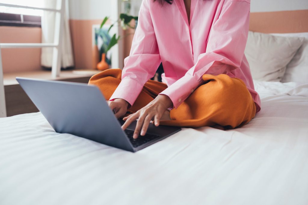 A woman sitting on a bed and typing on a laptop