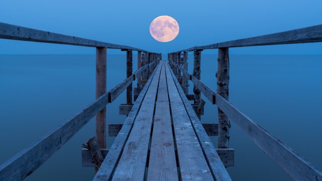 A full moon rising over a lake with a view down a long dock