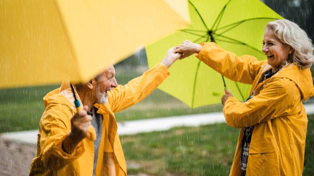 Happy senior couple in raincoats having fun while dancing with umbrellas during rainy day at the park.