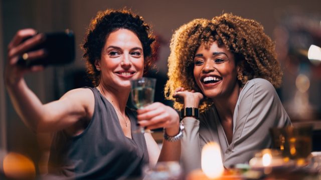 Cheerful smiling woman taking photography on a mobile phone with her friend during a dinner party.