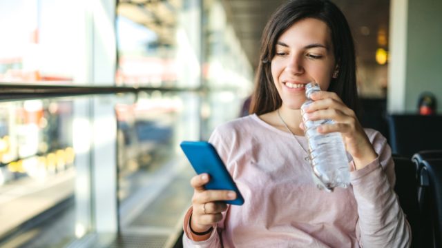 Young woman is drinking bottled water and using phone at the airport departure area