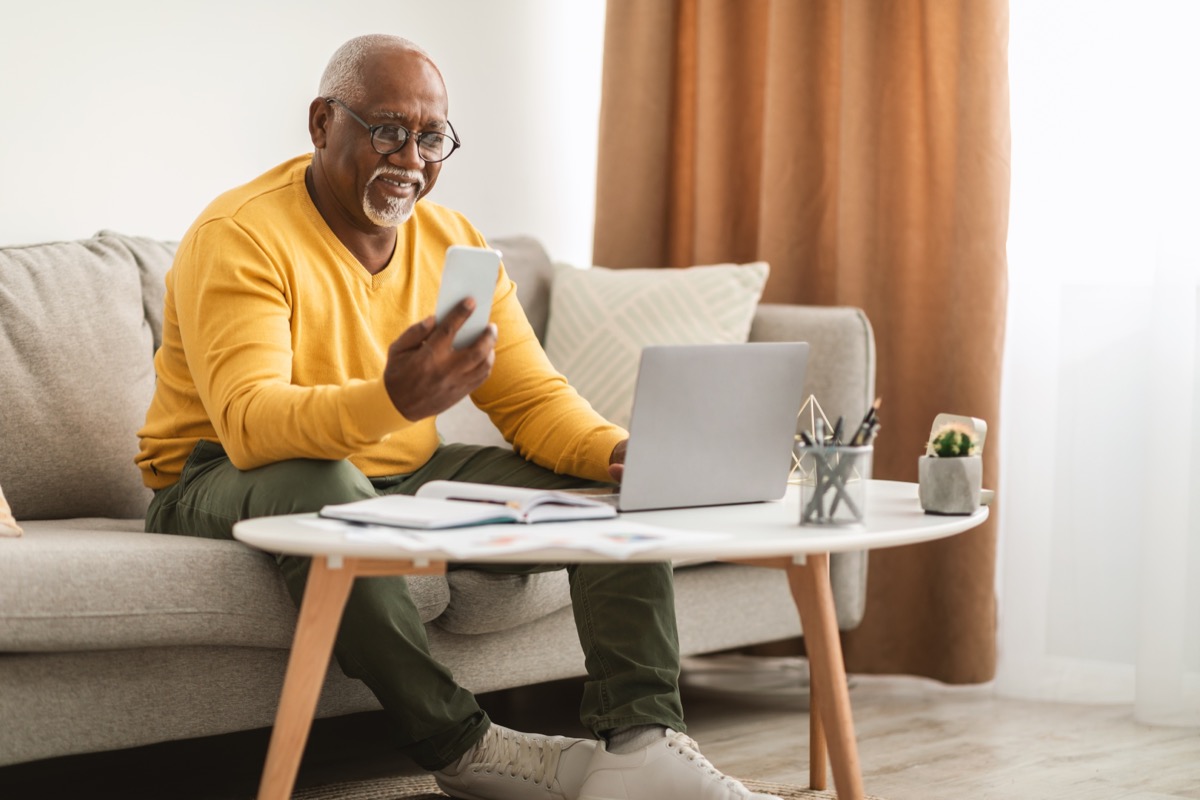 man in a yellow sweater smiling as he looks at his phone while sitting on the couch
