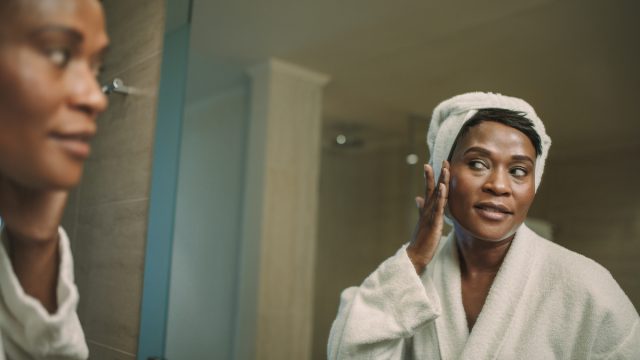 A mature woman wearing a white bathrobe and white hair towel stands in front of the mirror touching her face.