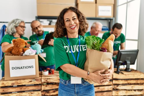 A smiling middle-aged woman wearing a green "volunteer" t-shirt holds a bag of food to be donated.