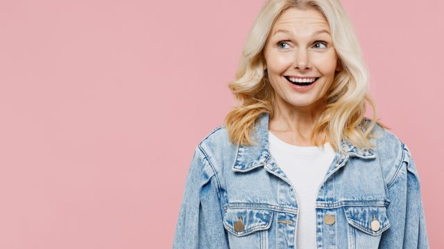 Elderly surprised amazed fun happy woman 50s wearing denim jacket look aside on workspace area mock up isolated on plain pastel light pink background studio portrait. People lifestyle fashion concept